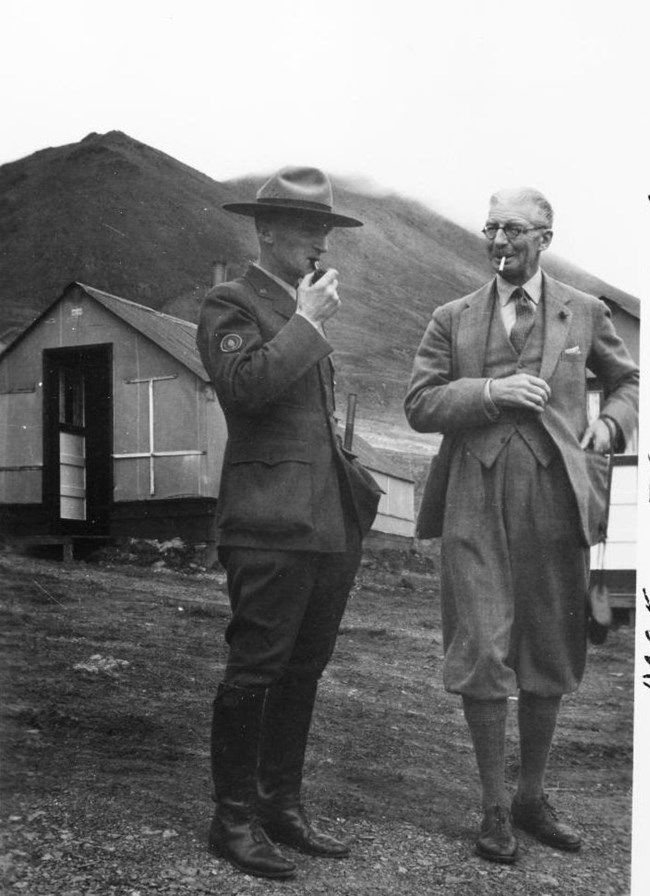 a park ranger and another man standing near a small cabin