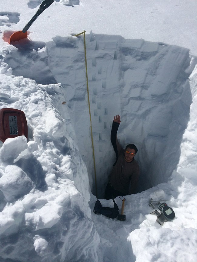 man waves from 10 feet down inside a hole dug into deep snow, with red snow shovel standing upright on the snow surface above