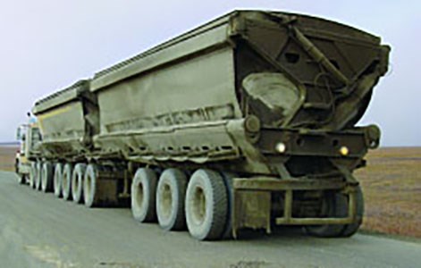 Semi-truck with two trailers hauling ore on a dusty, gravel road.