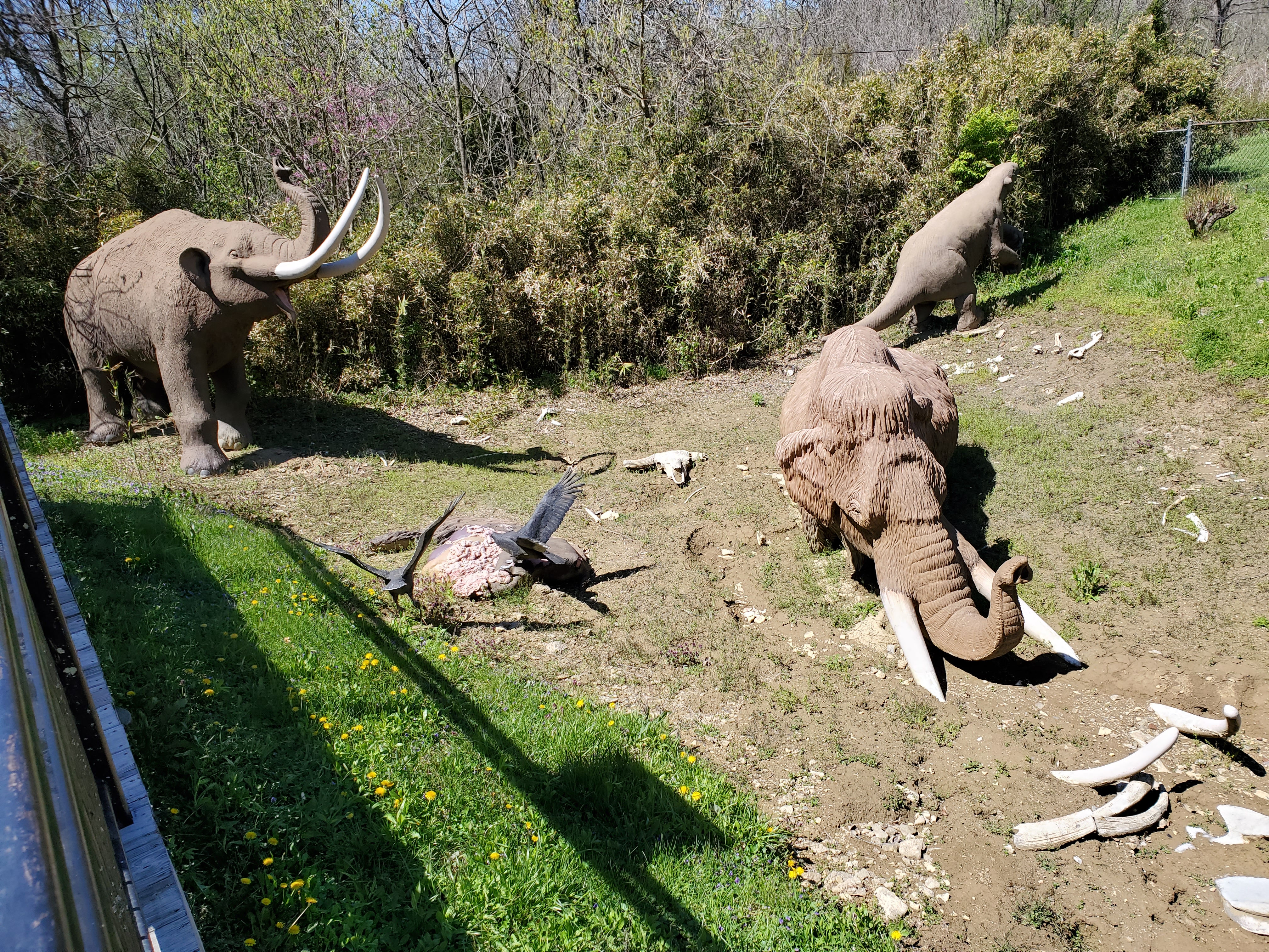 Models of a mastodon, mammoth, sloth, and vultures amid scattered bones situated in a grassy field.