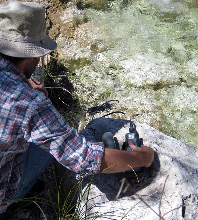 A person crouched down by flowing water holding water quality equipment.
