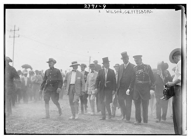 Several men walking side by side. Two men wear top hats and two are in uniform.