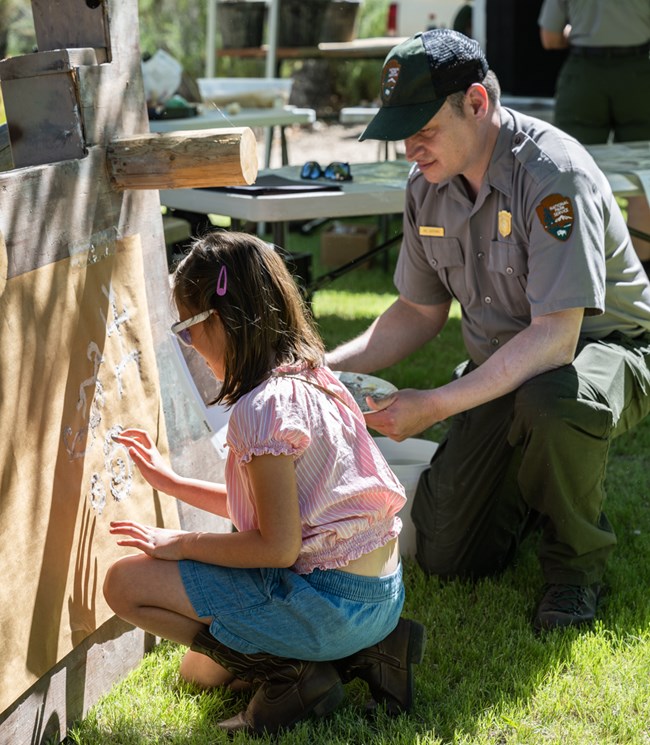 A ranger kneeling down next to a child. The child is finger painting with clay onto a brown sheet of paper. The activity is about creating your own pictograph.