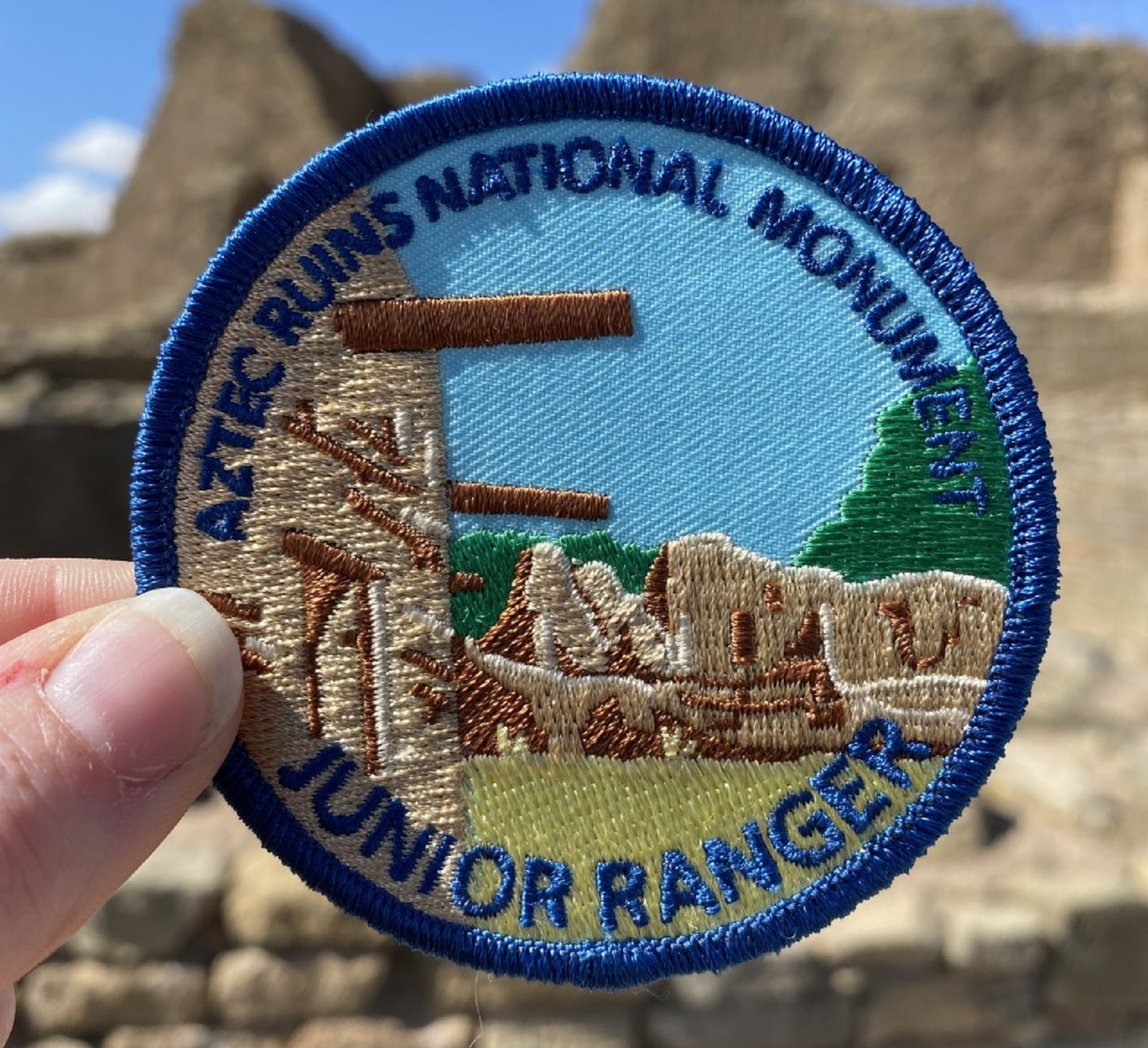 A hand holding a circular blue patch that says Aztec Ruins National Monument Junior Ranger