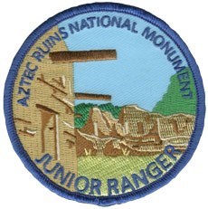 A blue circular patch that says Aztec Ruins National Monument Junior Ranger and shows a side of the Great Kiva and tan colored ruins in the background.