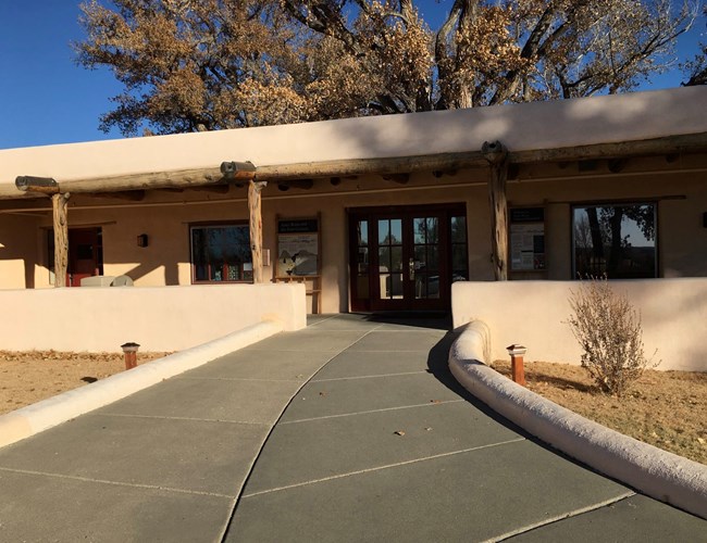 The front porch of the Aztec Ruins visitor center showing stucco walls, wooden columns, and a glass front door.