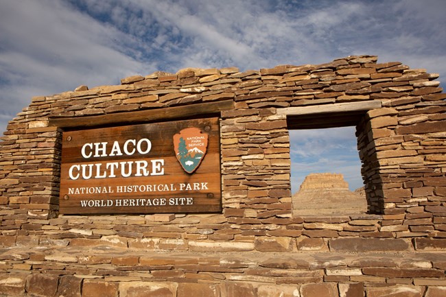 The entrance sign to Chaco Culture National Historical Park. It is made up of stacks of sandstone with a window on the right side showing a tall butte in the opening.