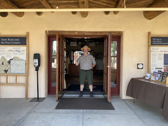 A ranger standing in the front doorway of the visitor center smiling and waving.