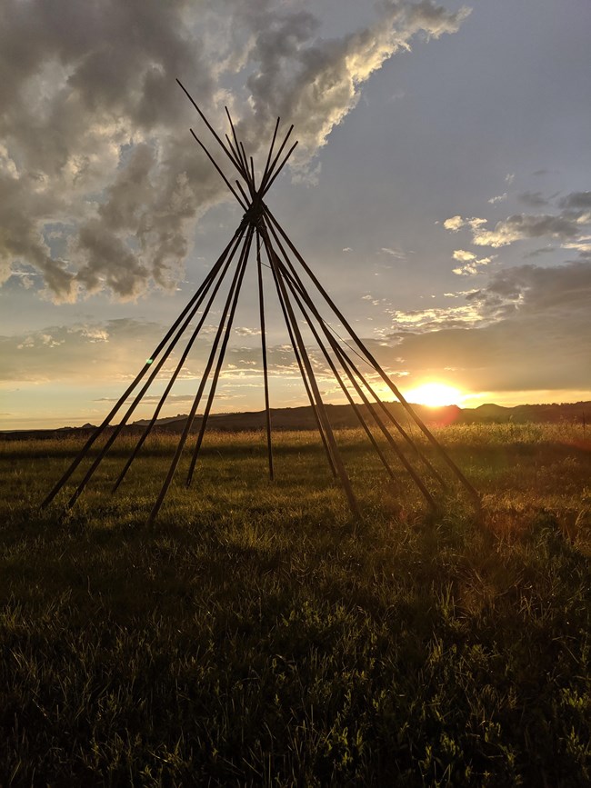 several long poles arranged in a tipi shape against a sunset and badlands buttes in background.