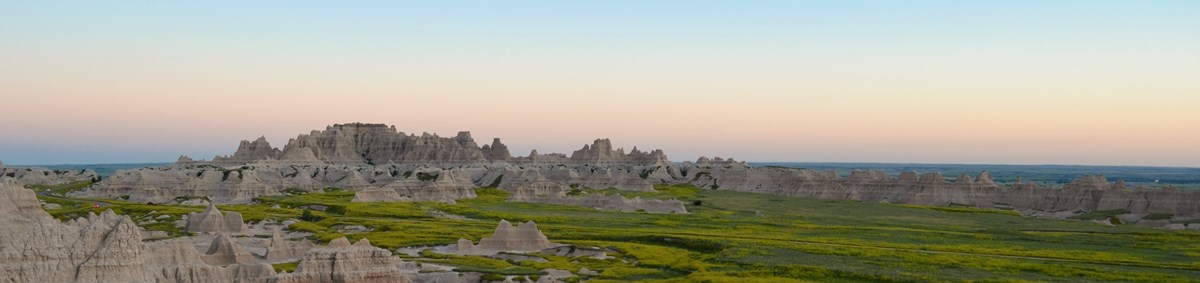 a pale sunset behind an expanse of badlands buttes and bright green prairie.