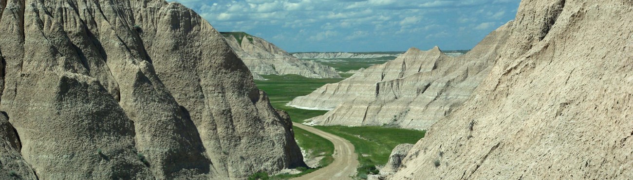 a dirt road winds between a pair of steep badlands buttes before disappearing behind the left butte, with a green grassy prairie in the distance.