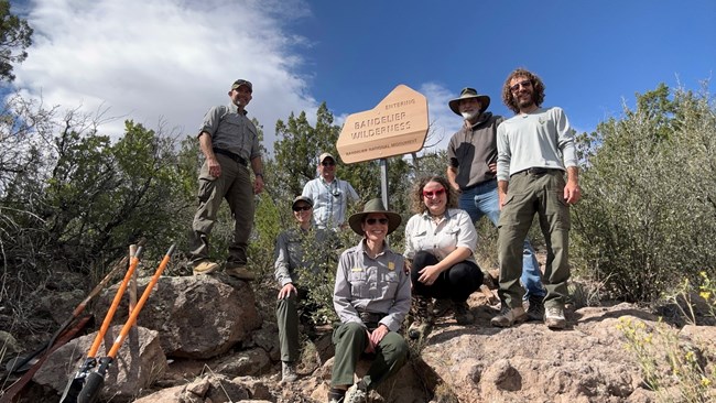 A group of men and women stand around a wooden sign that reads, "Entering Bandelier Wilderness, Bandelier National Monument."