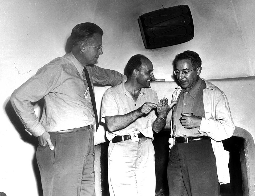 Three men stand and talk in front of a fireplace.