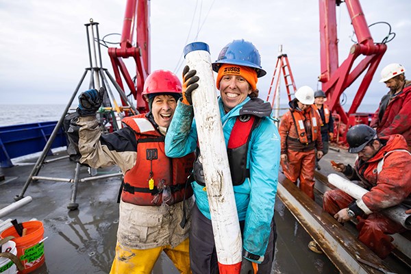Two women, both smiling, stand on the deck. The one on the right holds a long white tube.