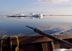 A seal hunting harpoon rests on the side of a wooden boat, looking out on a glassy sea with sea ice on the horizon