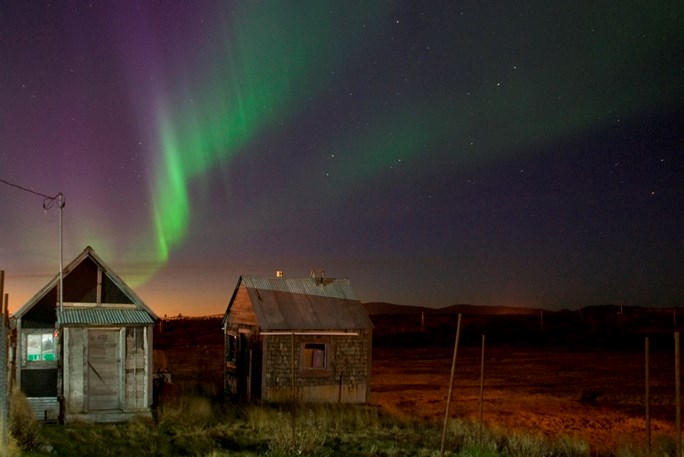 Purple and green northern lights hang like a curtain in the night sky over two wooden shacks in Nome