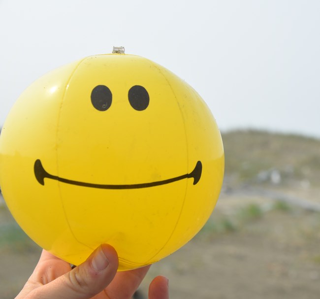 A hand holding up a yellow smiley-face ball, picked up as marine debris on the coast of Bering Land Bridge.