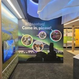 An exhibit panel with the northern lights, granite tors in the shape of a drummer, and photos of people featured in circles. Text reads "Itignaatuq! Come in, please!"