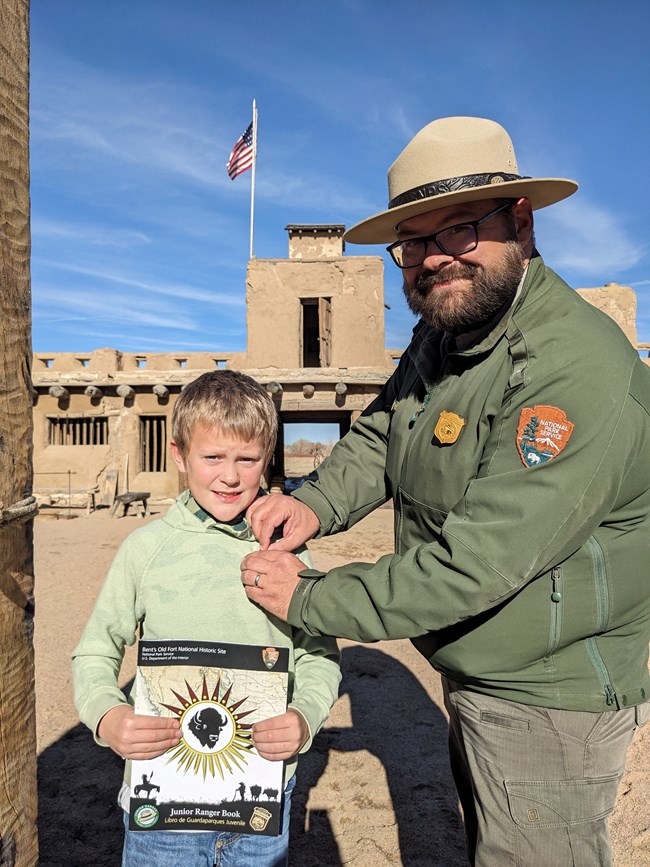 Photo of young boy holding a Junior Ranger book standing in fort plaza with Park Ranger pinning a badge on shirt.