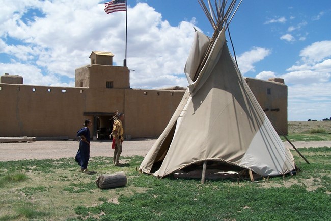Two people dressed in the historic costume of the Cheyenne stand by a tipi in front of the fort.