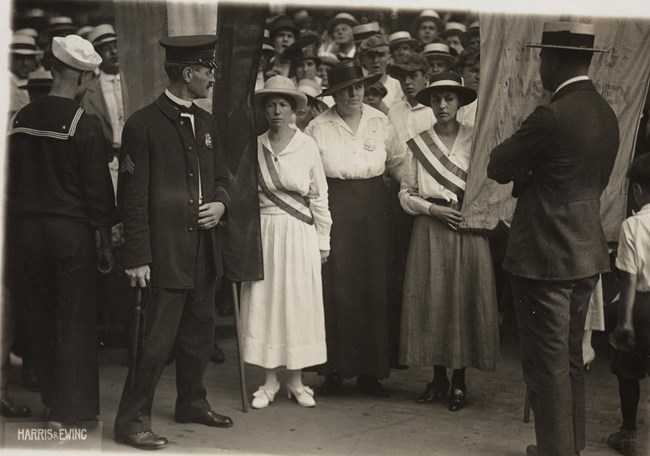 A policewoman holds the arms of two suffragists in a crowd of men
