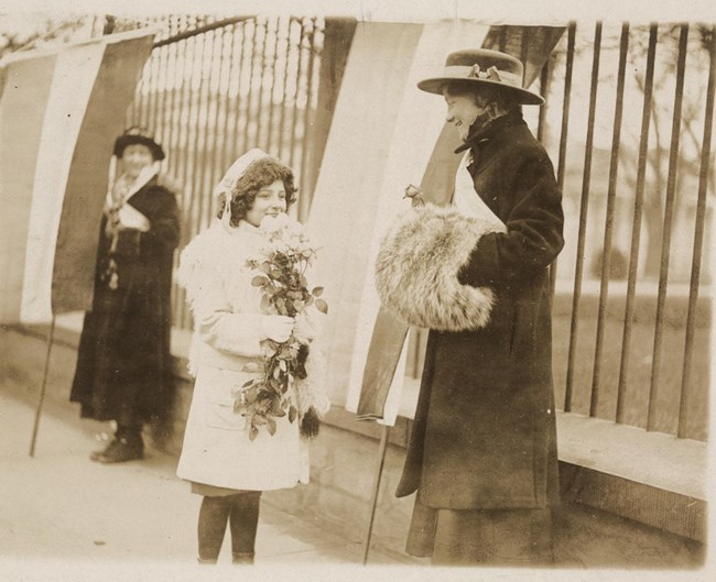 a young girl giving a bouquet of flowers to a smiling picket with fur muff and suffrage banner as she is picketing outside the White House while another woman holding a banner looks on