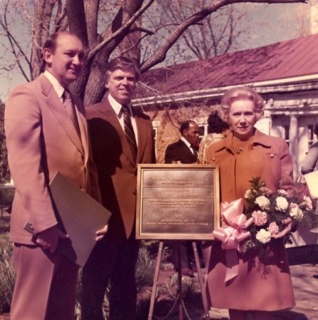 Two men and a woman holding flowers pose around National Historic Landmark plaque in NWP garden
