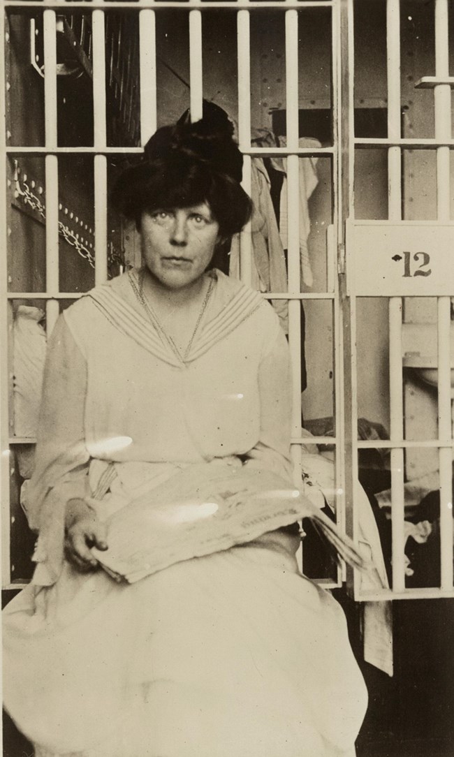 Lucy Burns sitting in front of a jail cell holding a paper in her lap