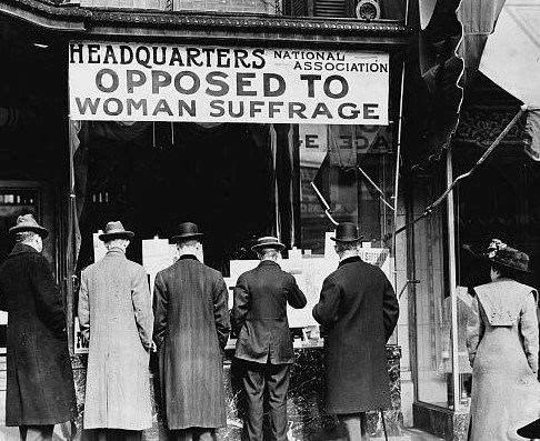 Photograph shows men looking at material posted in the window of the National Anti-Suffrage Association headquarters; sign in window reads "Headquarters National Association Opposed to Woman Suffrage".