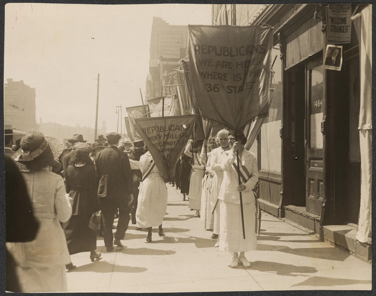 Photograph of line of suffragists (right) picketing with banners on city sidewalks, with passerby (left). Banner in foreground reads: "Republicans We Are Here Where is The 36th State?"