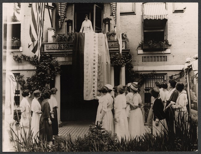 Alice Paul standing on balcony of headquarters with ratification banner unfurled and group of women gathered on the sidewalk below