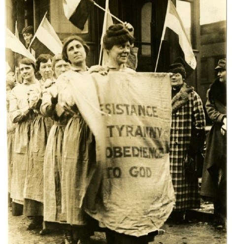 A line of women in prison dress in a train station. The first holds a banner reading "Resistance to Tyranny is Obedience to God"
