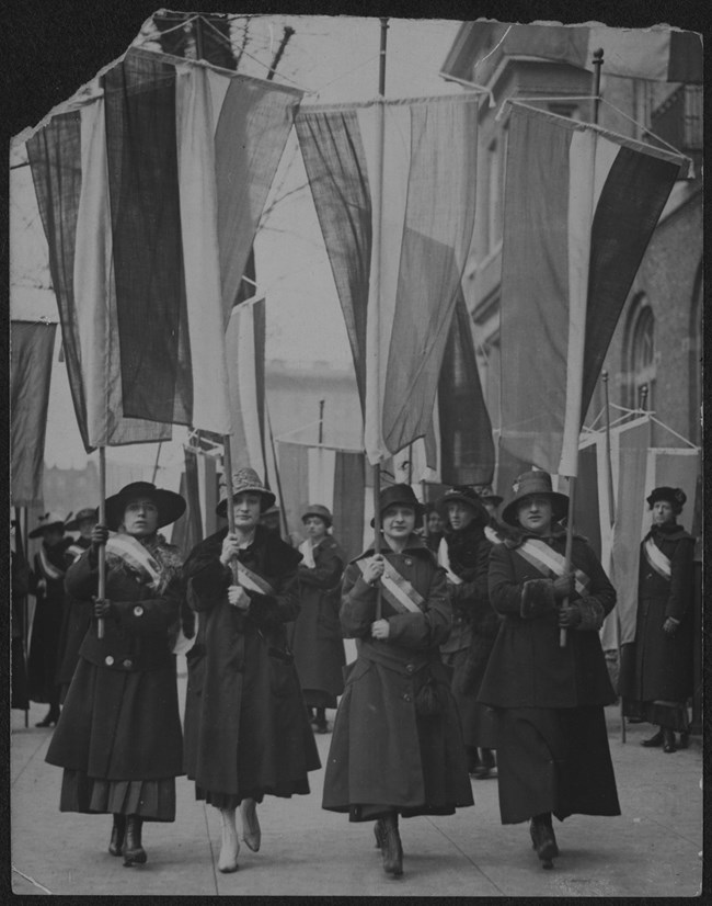 Four women wearing coats, hats, and suffrage sashes carrying tri-colored banners