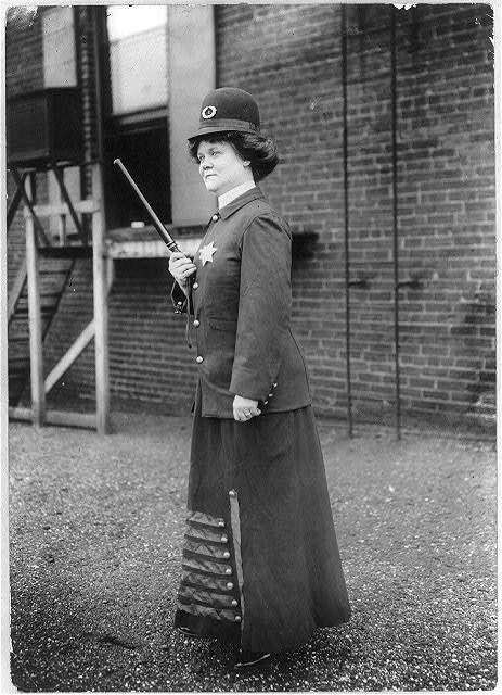 Woman posing in a police uniform with long skirt holding a billy club