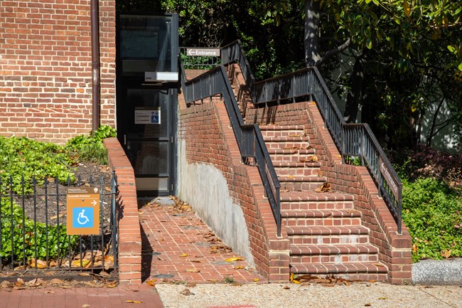 A brick walkway leading to the door of a wheelchair lift next to a brick staircase, both leading up to a brick building.
