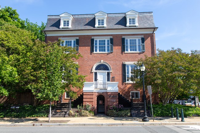 A four-story brick house with trees and a sidewalk out front and two staircases leading up to a white door.