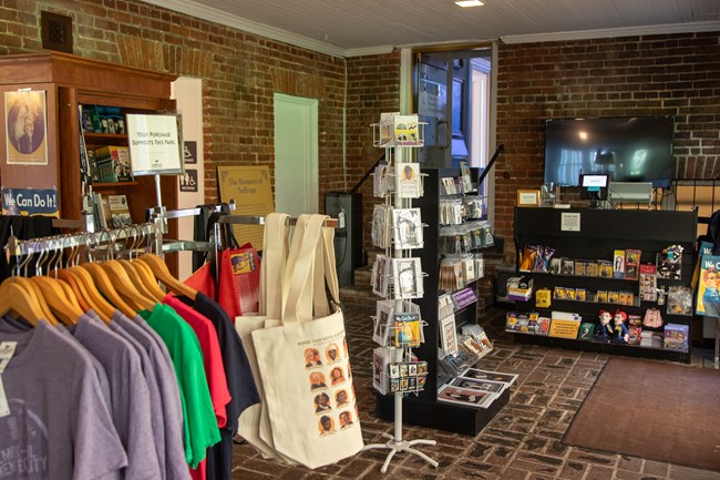 Racks of t-shirts, totes, postcards, and other souvenirs in a room with brick walls and floors.