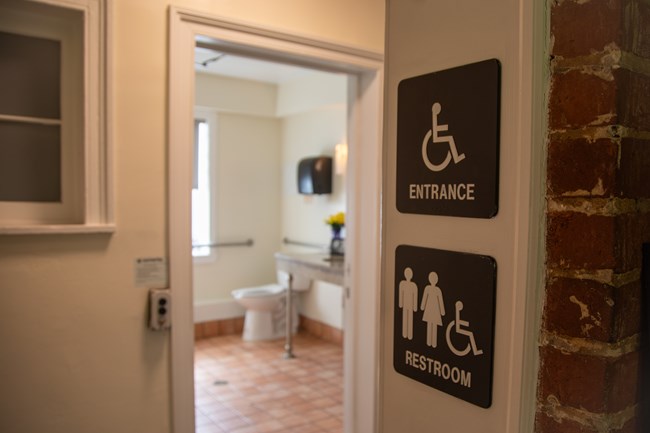 A doorway with signs for an accessible restroom and accessible entrance with a restroom in the background.