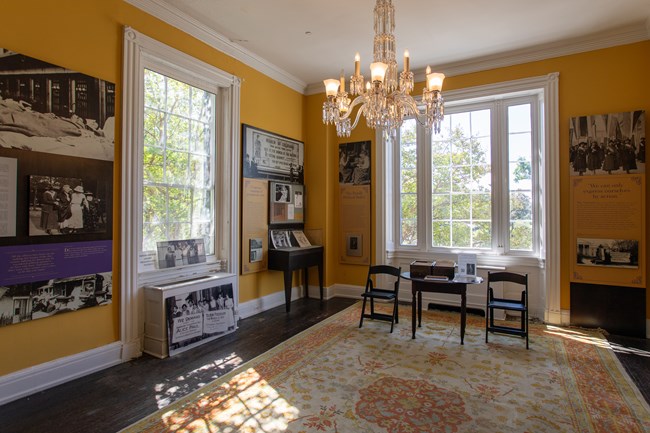 A room with high ceilings and big windows, a rug, chairs, and exhibits with photos and text hung on the walls.