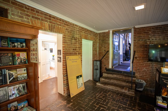 A room with brick floors and two doorways, one leading into a hallway and another with four steps up to a decorated foyer.
