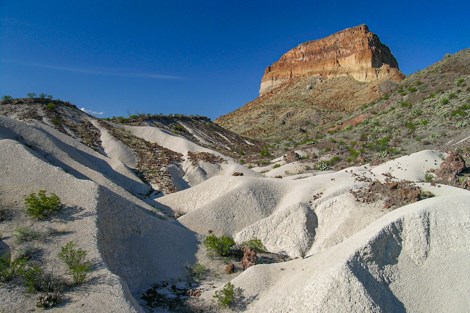 White volcanic ash dominates the foreground of the scene, with a high peak of red, orange and yellow rock layers standing above.