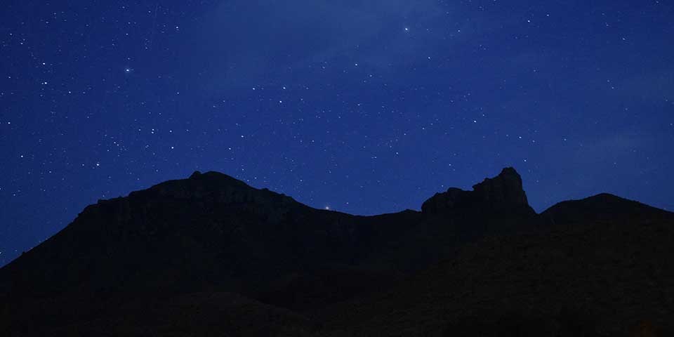 Bright stars and wisps of galactic dust glow in a deep blue night sky over silhouettes of mountains.