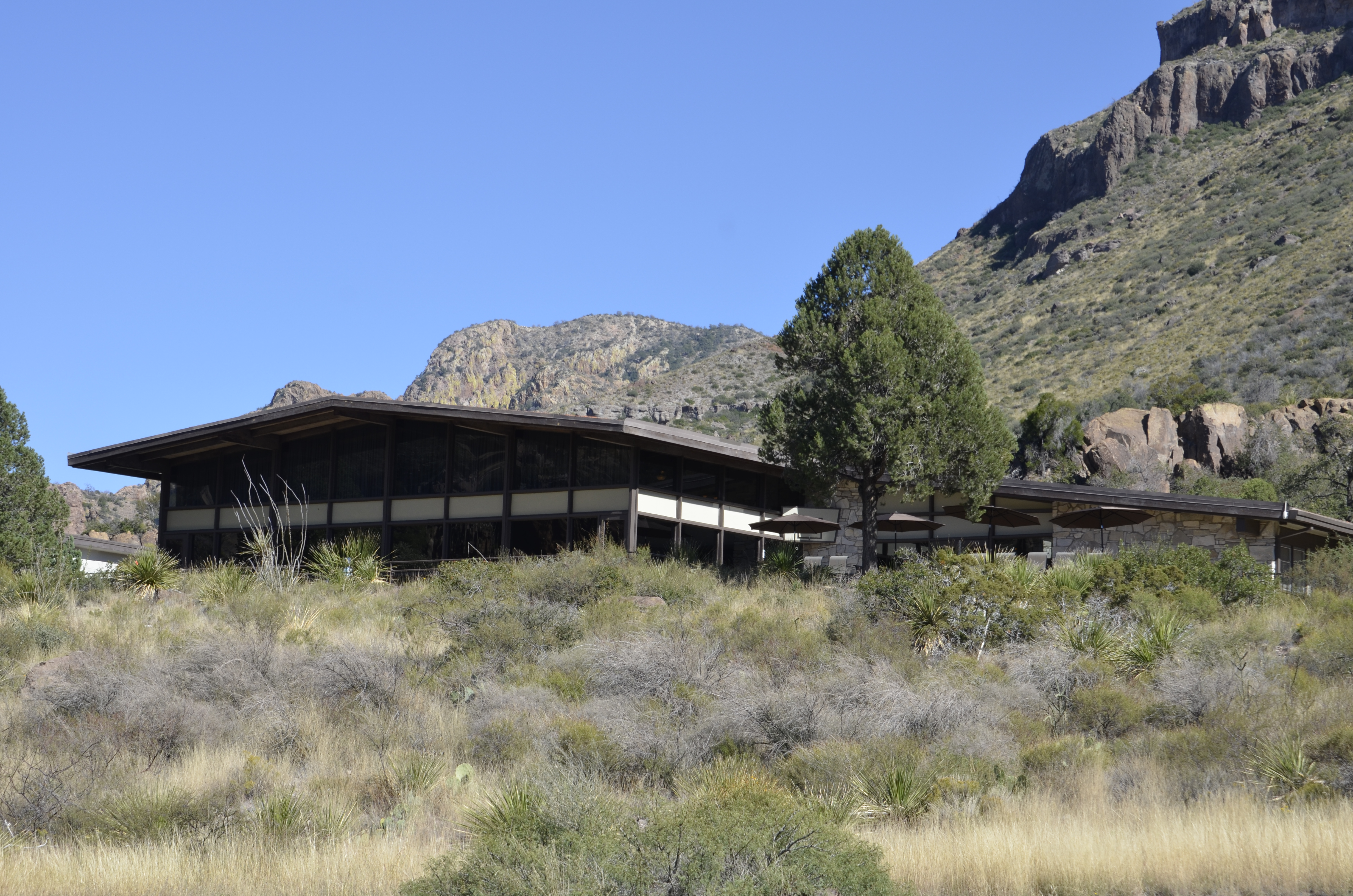 A brown building sits on a low hill with moutains rising behind it.