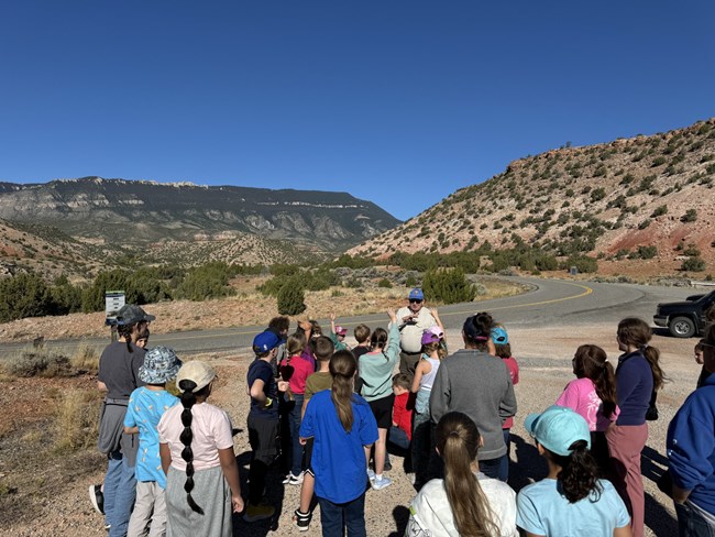 A group of students gathered around a park volunteer with mountains in the background.