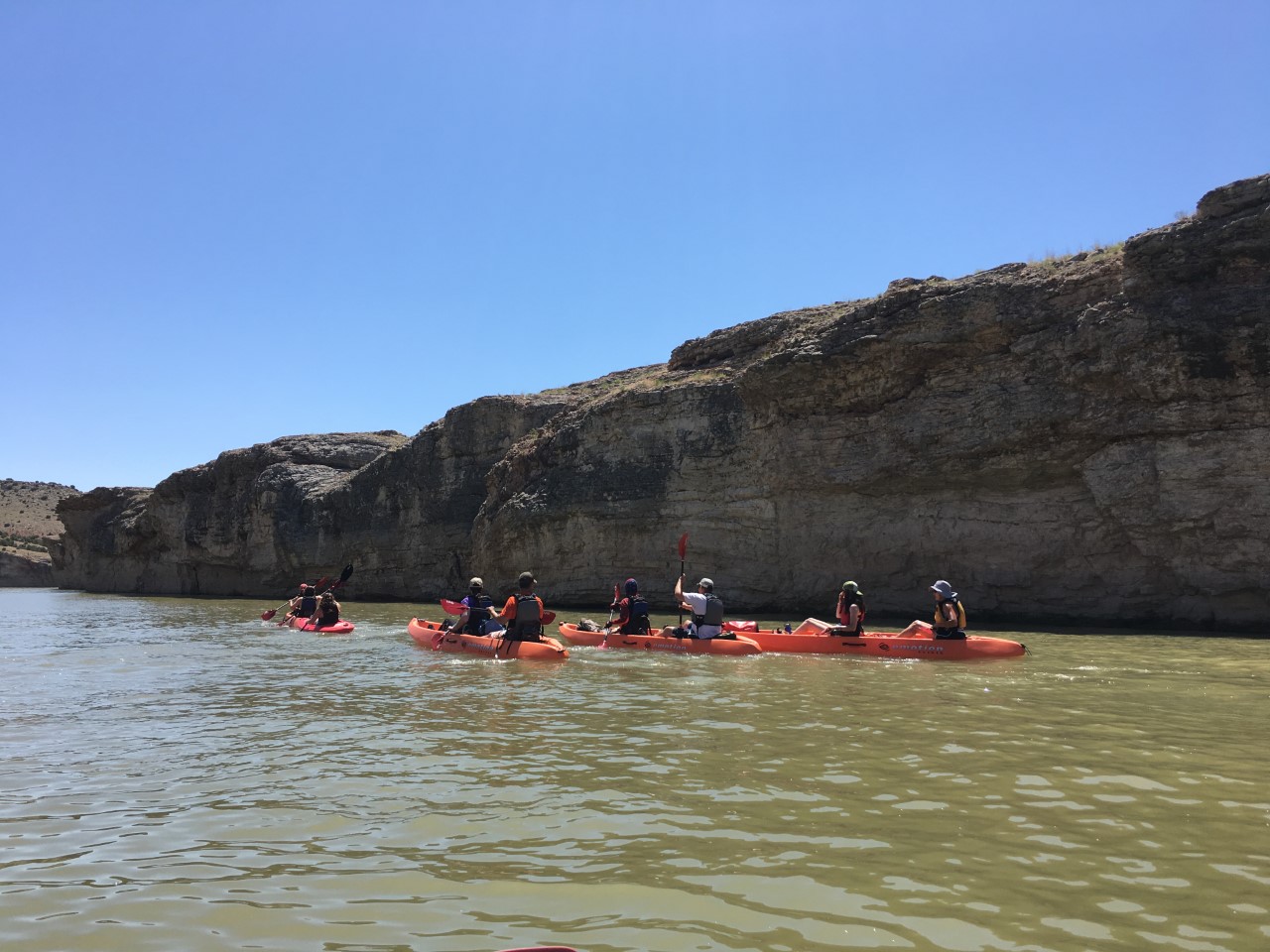 Several orange two person kayaks full of paddlers along the cliff.