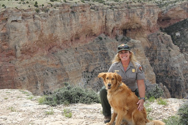 Park Ranger kneeling next to Golden Retriever with limestone canyon wall in the background.