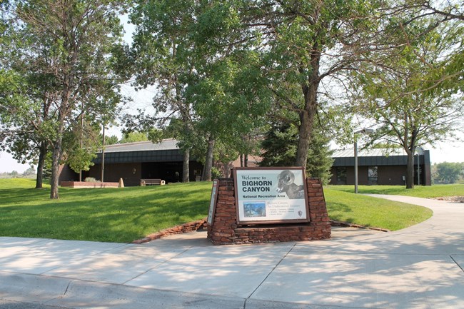 Welcome to Bighorn Canyon National Recreation Area sign in front of trees, a grassy lawn, and a brown building.