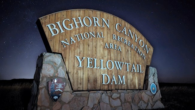 A lit up wooden and stone sign reading, "Bighorn Canyon National Recreation Area Yellowtail Dam" with a night sky full of stars in the background.