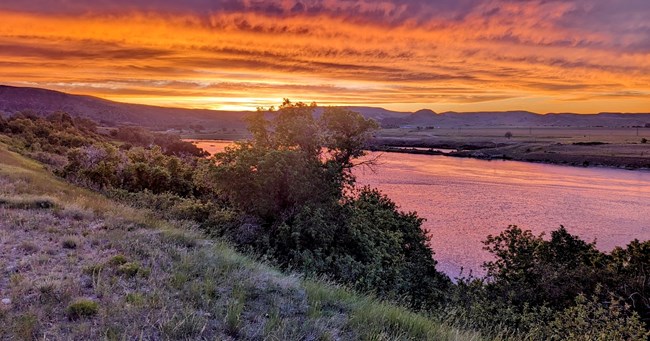 An orange and purple sunset reflecting onto the water of the Bighorn River. trees and grass in the foregroud with rolling hills in the background.