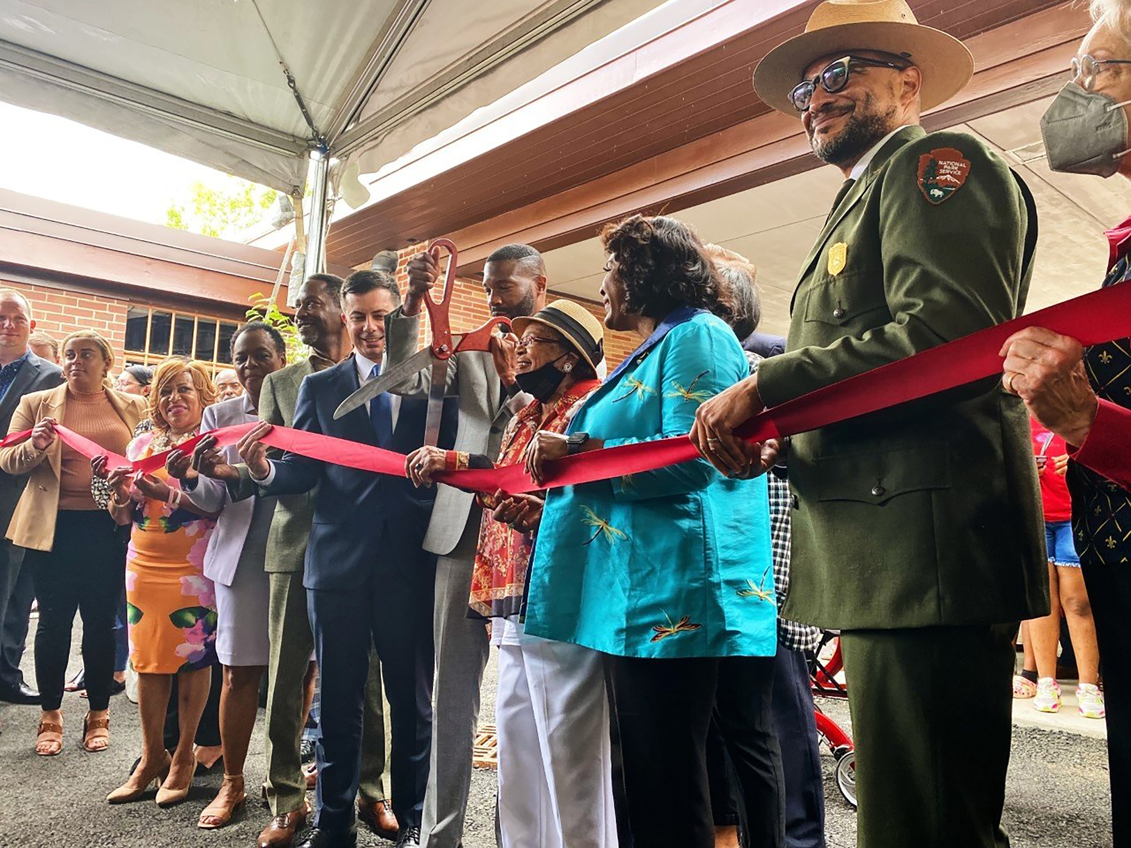 A line of people stands behind a ribbon. A man in a suit in the middle holds large scissors about to cut the ribbon.